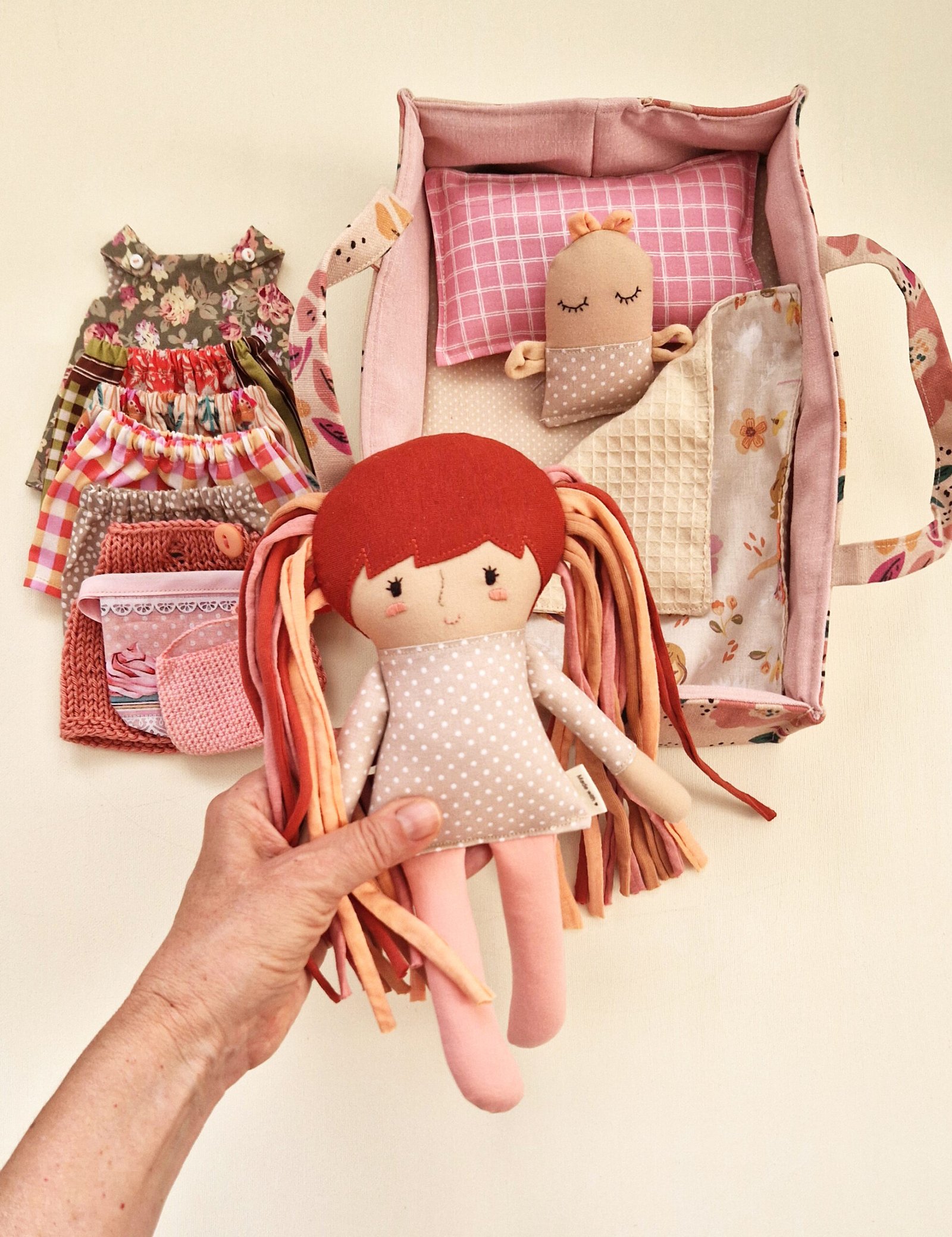 Fabric doll with long hair in pink and red tones displayed beside clothes and a fabric bag.