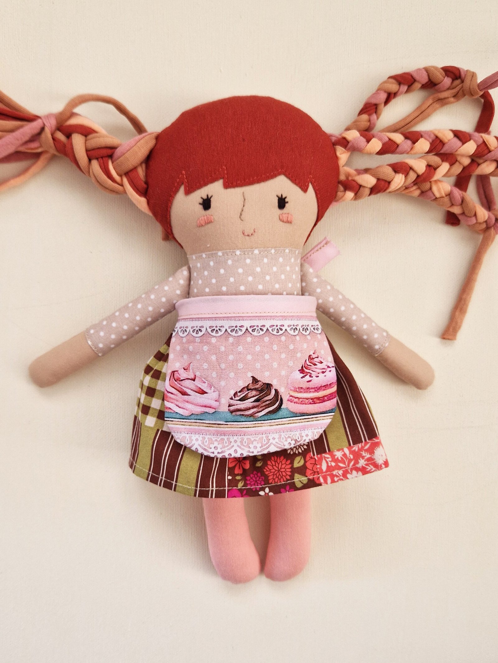 Fabric doll with long hair in pink and red tones displayed beside clothes and a fabric bag.