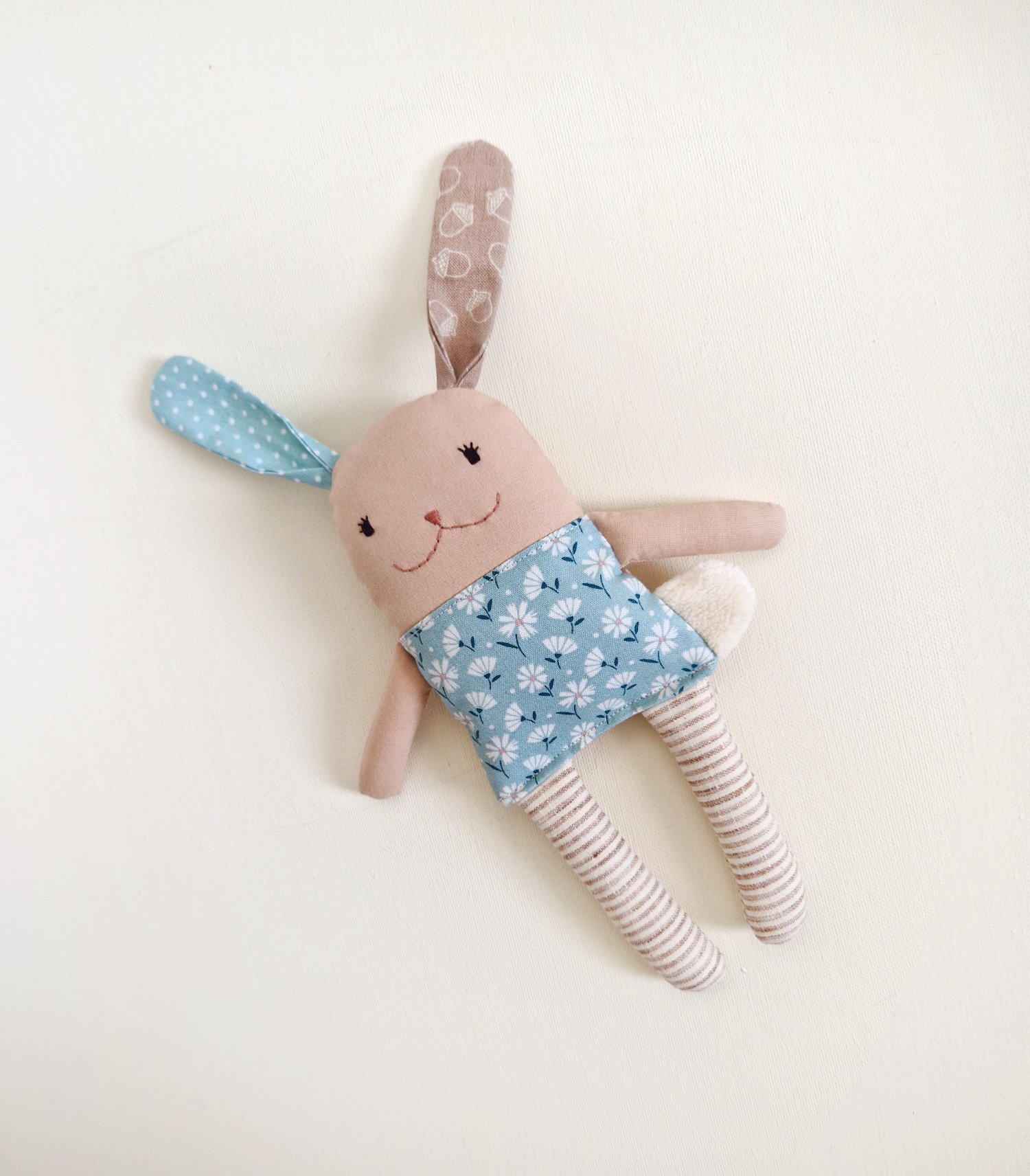 Front view of a handmade reversible fabric bunny plush toy for babies, with open embroidered eyes, a blue floral pattern shirt, and striped legs.