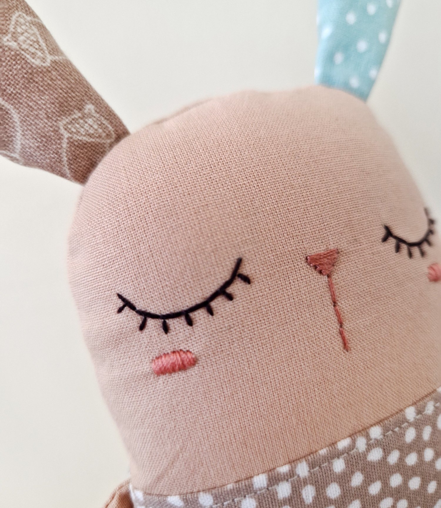 A soft textile baby toy shaped like a rabbit, featuring long mismatched ears (one blue polka dot, one brown with acorns) and a friendly stitched face.